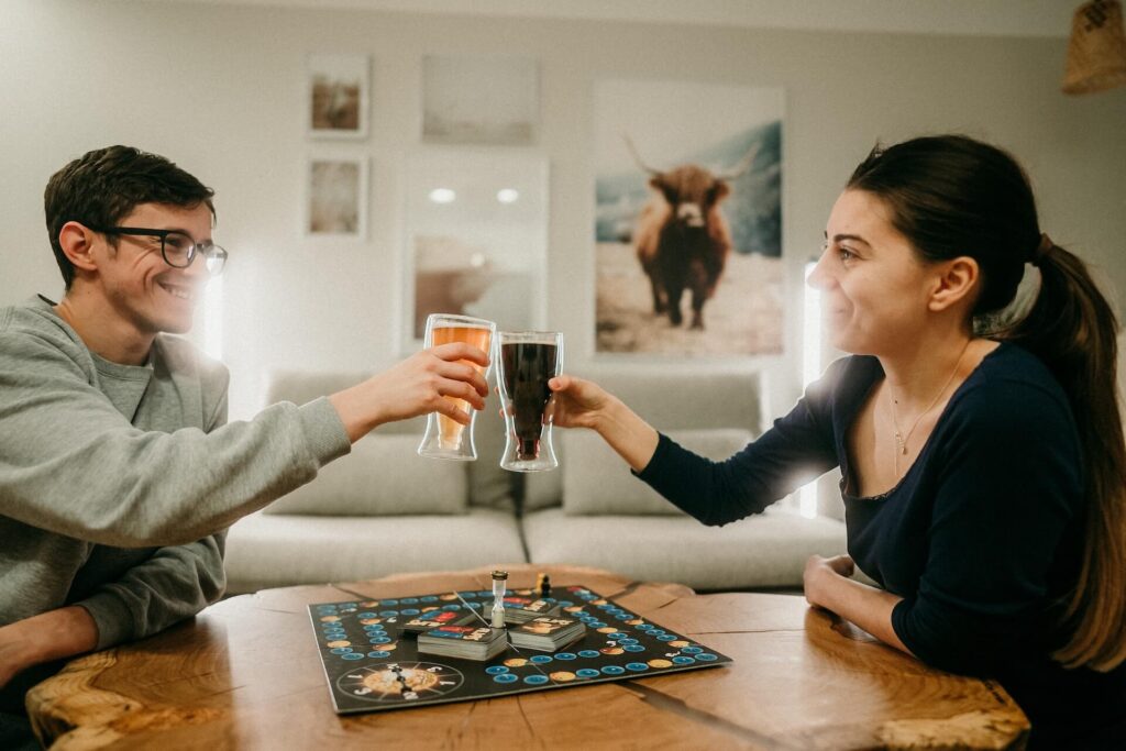 Couple toasting each other across a wooden table with a board game on it | Featured Image for the Best Board Games for Date Night Blog.
