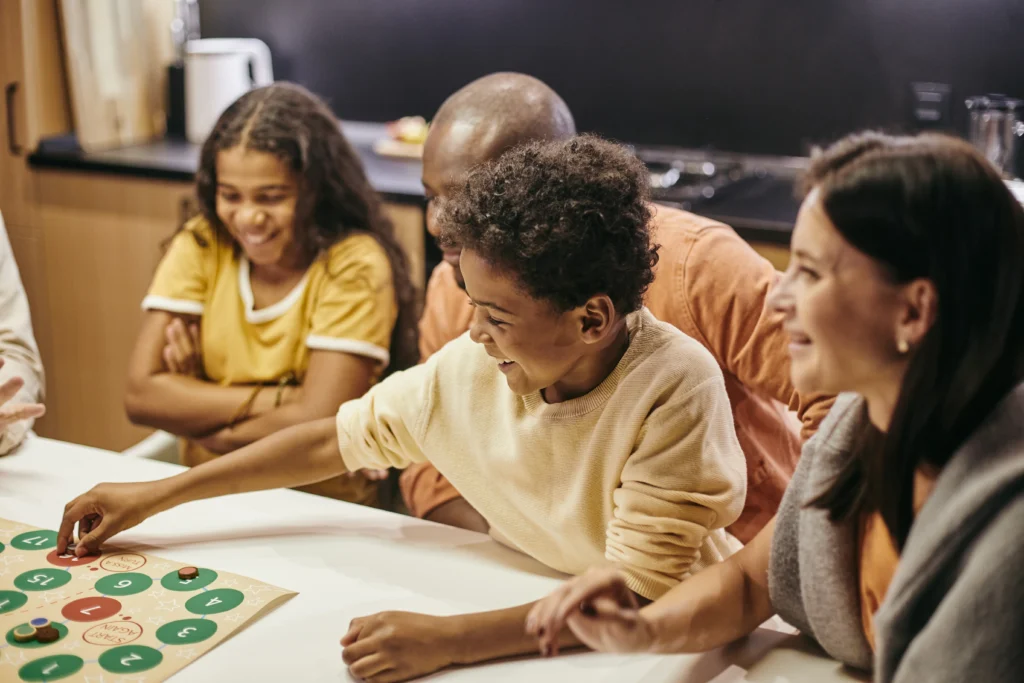 A happy group is gathered around a game. A child in a pale yellow sweater is about to move their game piece.