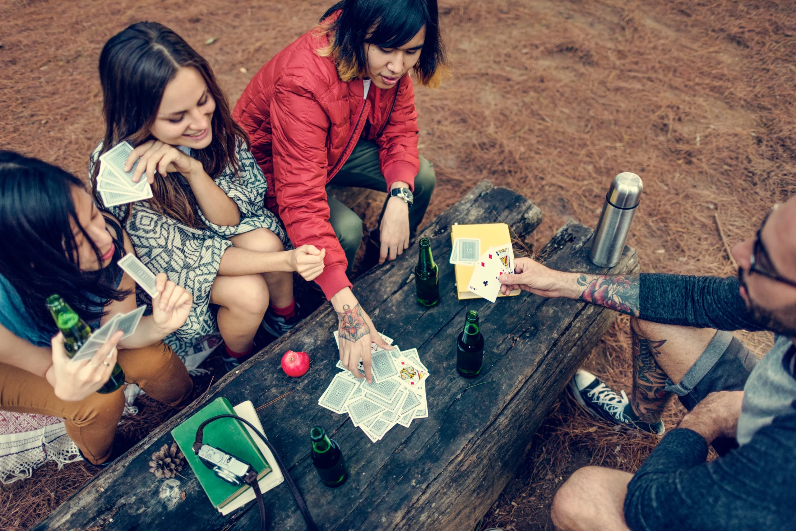 Four adults sit around a rustic, dark wood picnic table outdoors in a dry forest. The table is covered in playing cards.