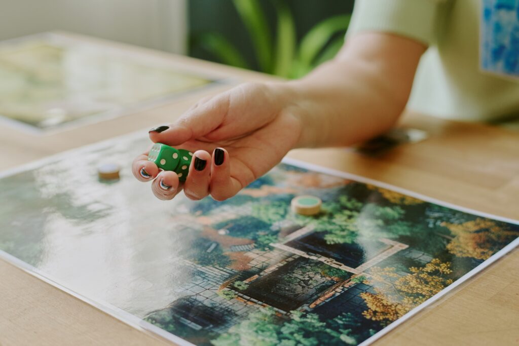 A hand with black nail polish holds a green dice above a board game on a wooden table. The game board shows a landscape of greens and yellows.