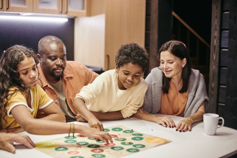 A family of four, including two adults and two children, are gathered around a white table playing a colorful board game