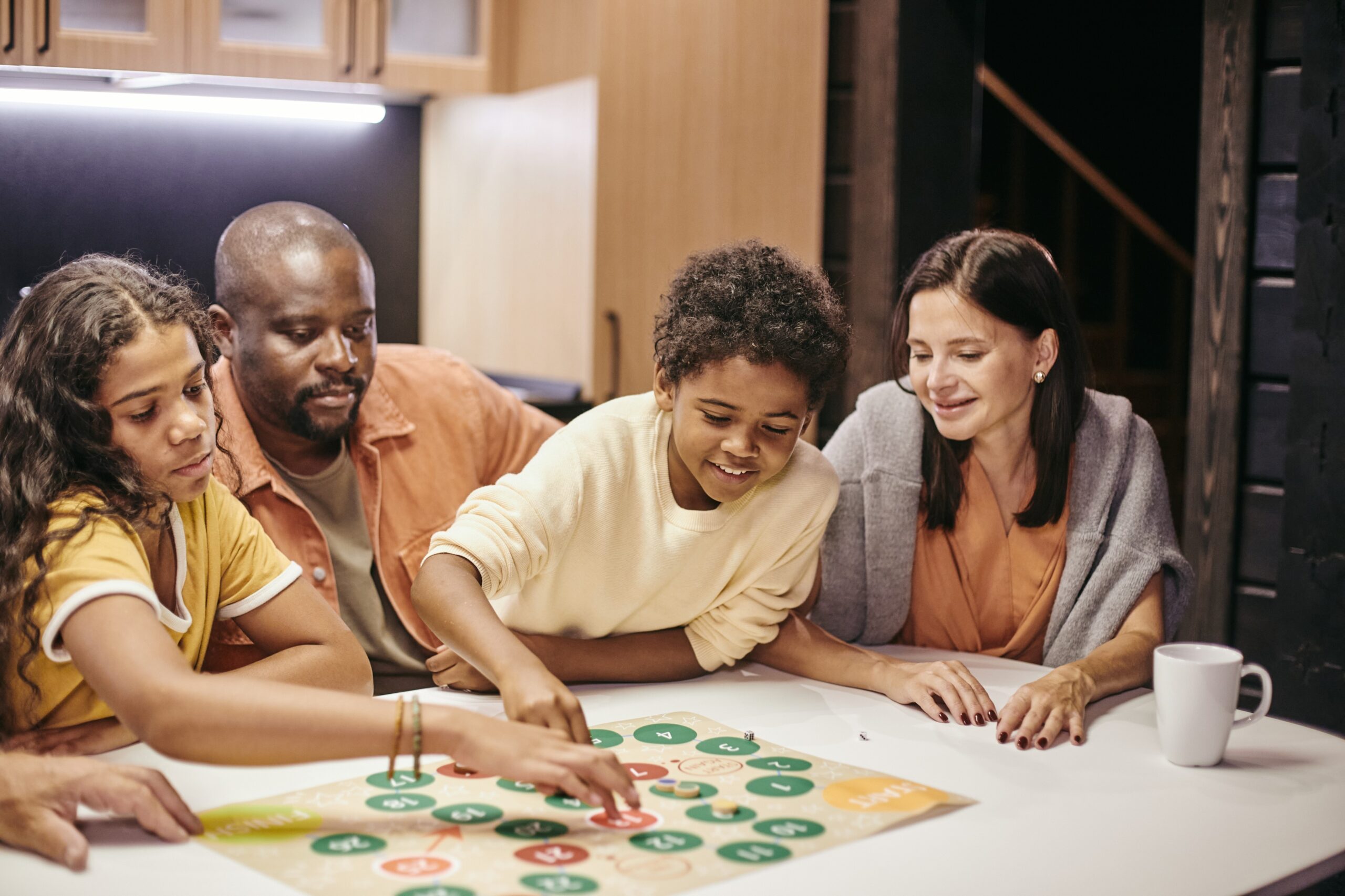 A family of four, including two adults and two children, are gathered around a white table playing a colorful board game